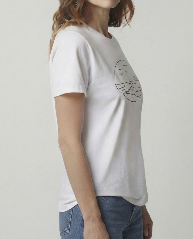 A woman wearing a white short-sleeve crewneck t-shirt with a black circular graphic of seascape on the chest, paired with blue jeans, standing against a light gray studio background.